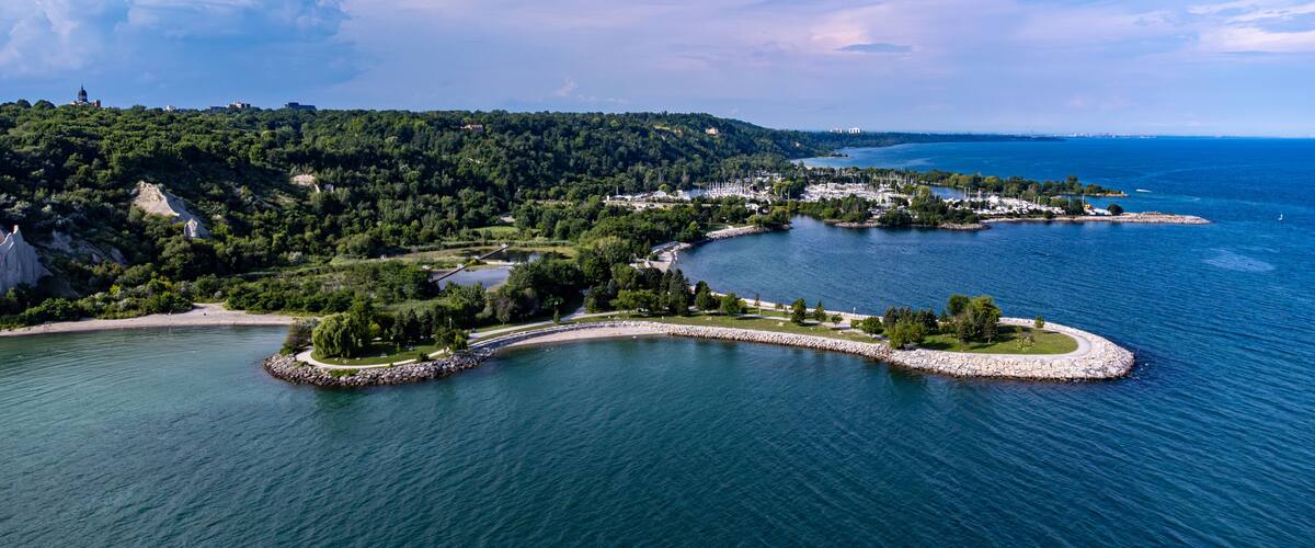 A coastal landscape with a marina, a curved breakwater, and a forested hill.