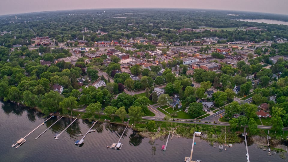 An aerial view of a lakeside town with dense greenery, a marina with boats, and a mix of residential and commercial buildings.