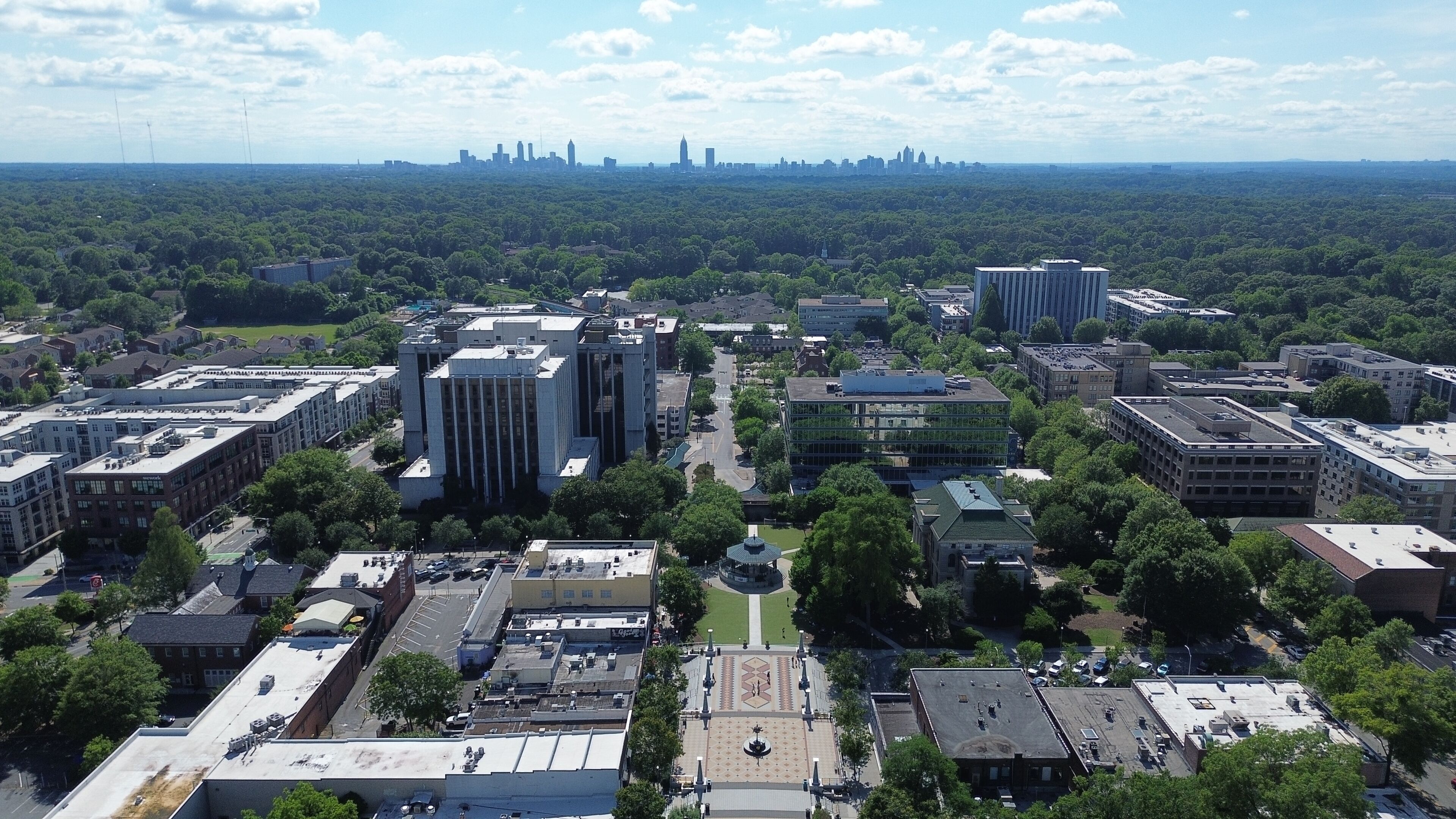 An aerial view of a city with a mix of modern and older buildings, surrounded by green spaces and a distant skyline.