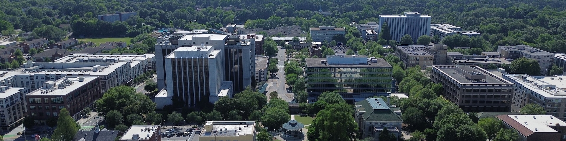 An aerial view of a city with a mix of modern and older buildings, surrounded by green spaces and a distant skyline.