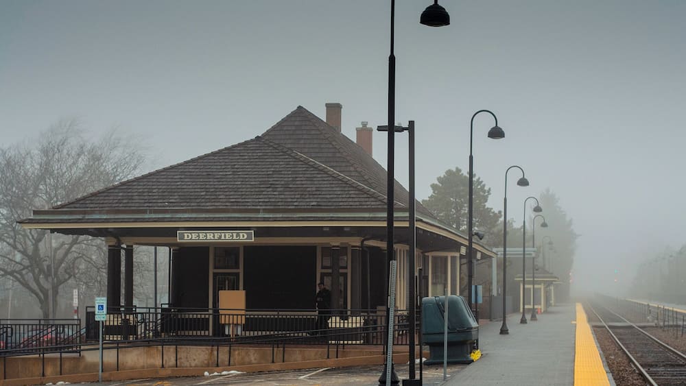 Ein nebliger Bahnhof namens 'DEERFIELD' mit überdachtem Wartebereich, Bahnsteig und Gleisen.