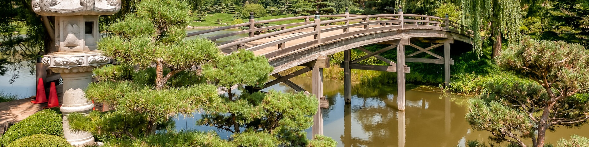 Un puente de madera sobre un estanque, rodeado de vegetación exuberante y un farol de piedra tradicional.