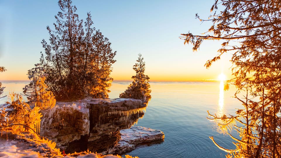 A sunset over a calm lake with trees and rocky outcrops.