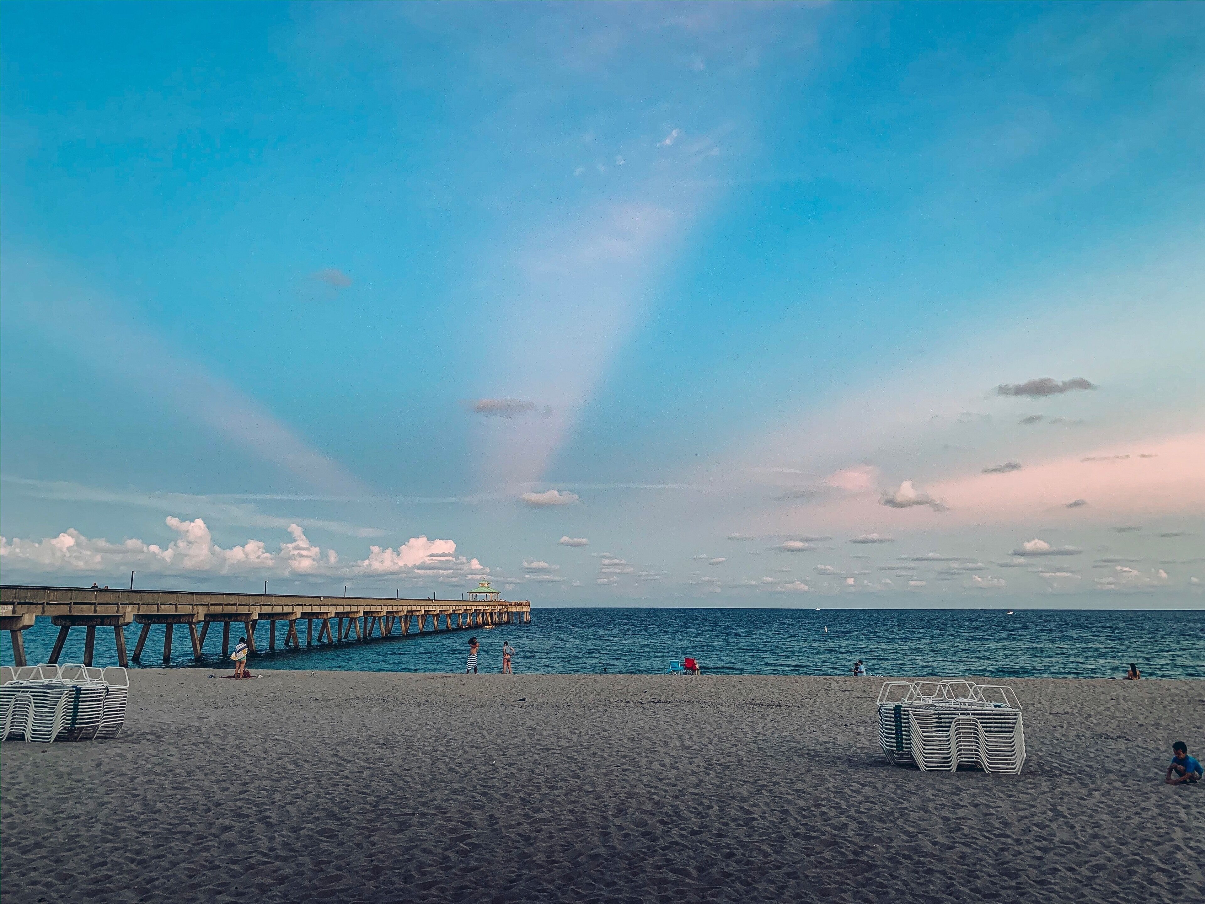 A beach with a pier extending into the water, people relaxing, and beach chairs stacked on the sand.
