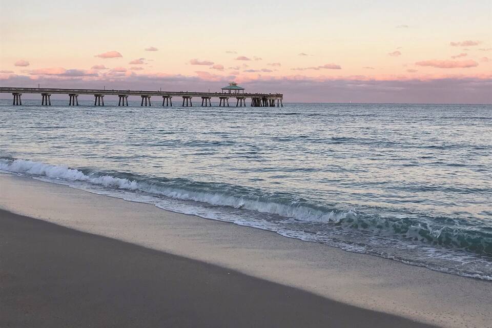 A beach with waves at sunset, a pier extending into the water, and a small structure on the pier.