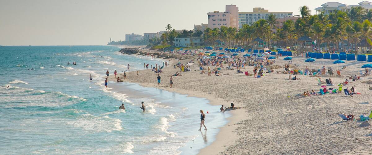 A crowded beach with people sunbathing and swimming, palm trees, and buildings in the background.