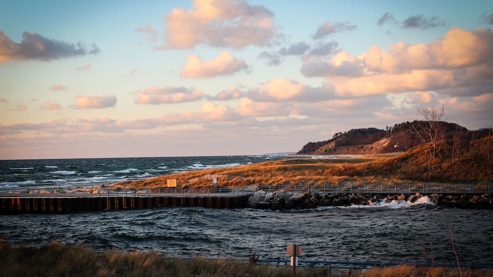 A coastal landscape with a pier extending into the sea, grassy dunes, and a hill with sparse vegetation.