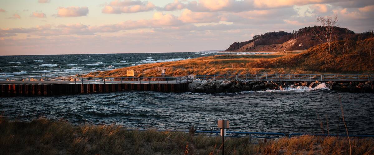 A coastal landscape with a pier extending into the sea, grassy dunes, and a hill with sparse vegetation.