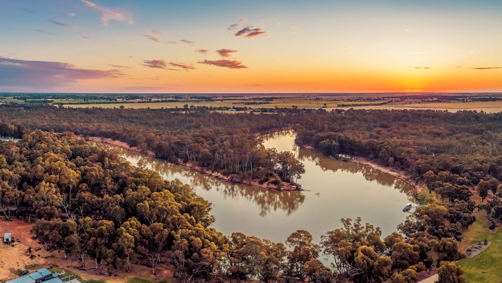 Ein Fluss schlängelt sich durch ein bewaldetes Gebiet, im Hintergrund Häuser und ein Sonnenuntergang.