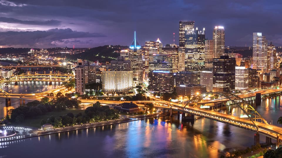 A nighttime cityscape with illuminated skyscrapers, a river, and a bridge.
