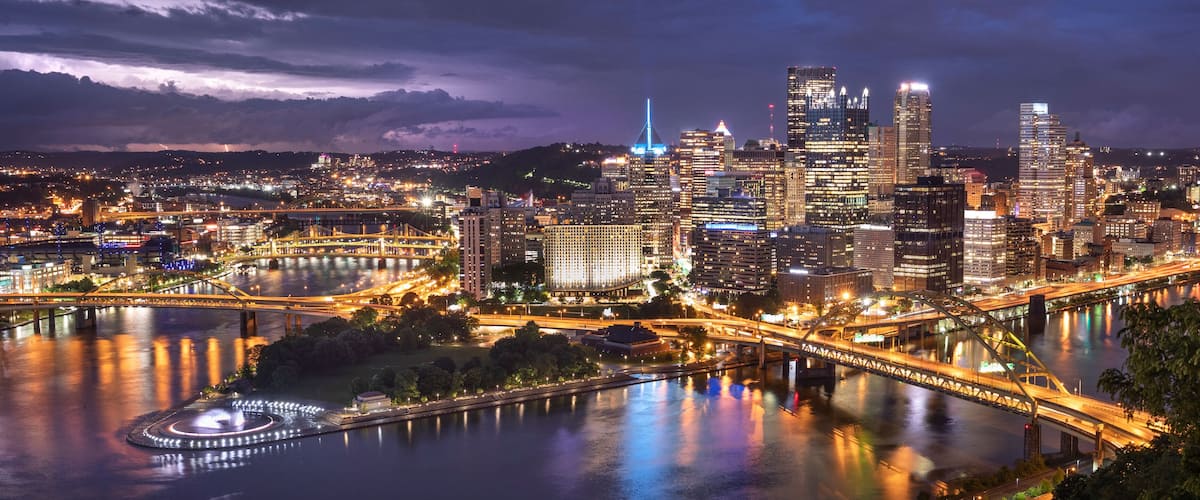A nighttime cityscape with illuminated skyscrapers, a river, and a bridge.