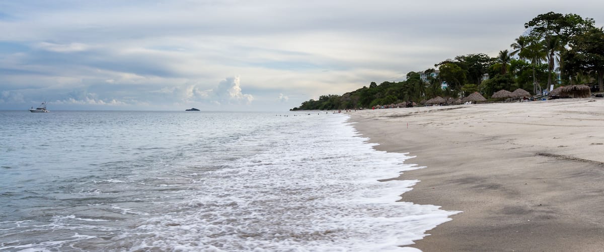 A beach with waves, a boat, and palm trees.