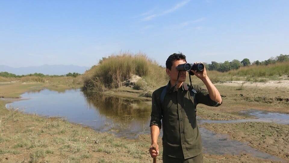 A person holding binoculars in a natural landscape with a body of water and dry grass.