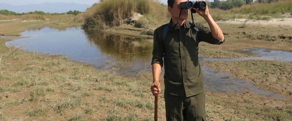 A person holding binoculars in a natural landscape with a body of water and dry grass.