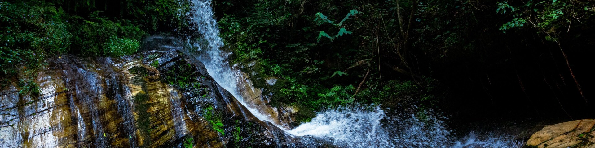 View of water cascading down a rugged, multi-colored rock face amidst verdant foliage, creating a vibrant contrast of textures and tones, Efon Alaaye, Ekiti, Nigeria.