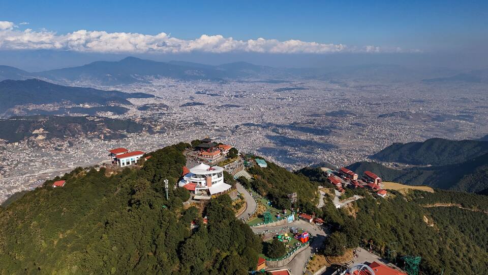 A mountainous landscape with a city in the valley below, a prominent white building with a red roof, and a large body of water in the distance.