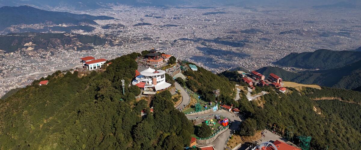 A mountainous landscape with a city in the valley below, a prominent white building with a red roof, and a large body of water in the distance.