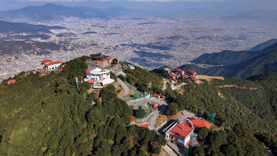 An aerial view of a mountainous area with a city in the background, a large white building with a red roof, and several smaller buildings with red roofs.