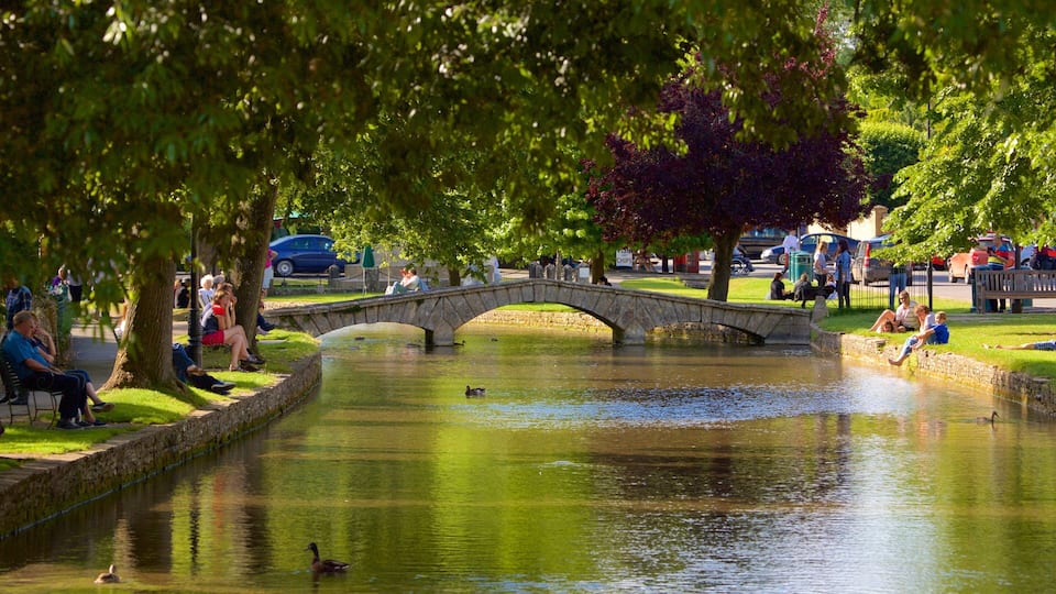 A stone bridge over a calm waterway in a park with people sitting on benches and ducks swimming.