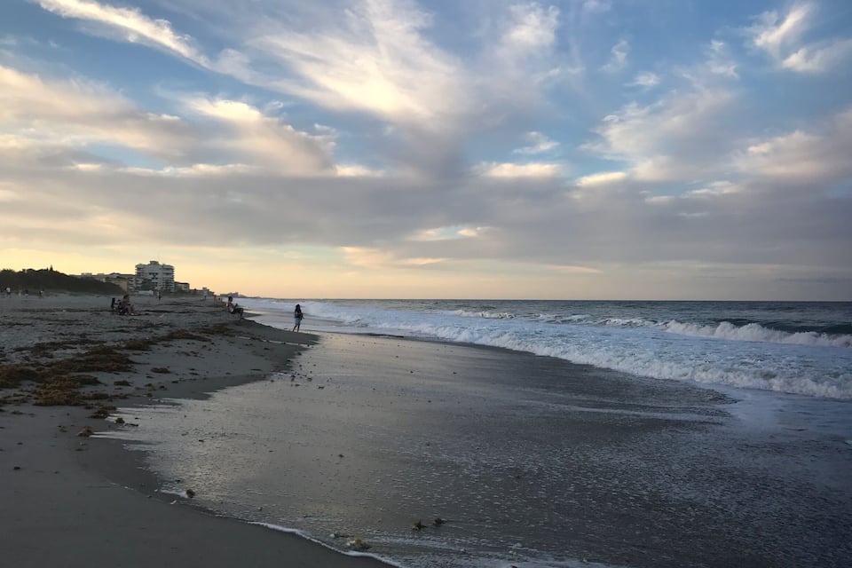 A beach with waves, a cloudy sky, and buildings in the background.