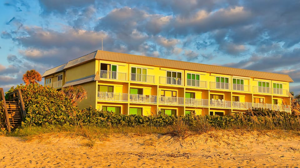 A yellow multi-story building with balconies, located on a sandy beach with vegetation.