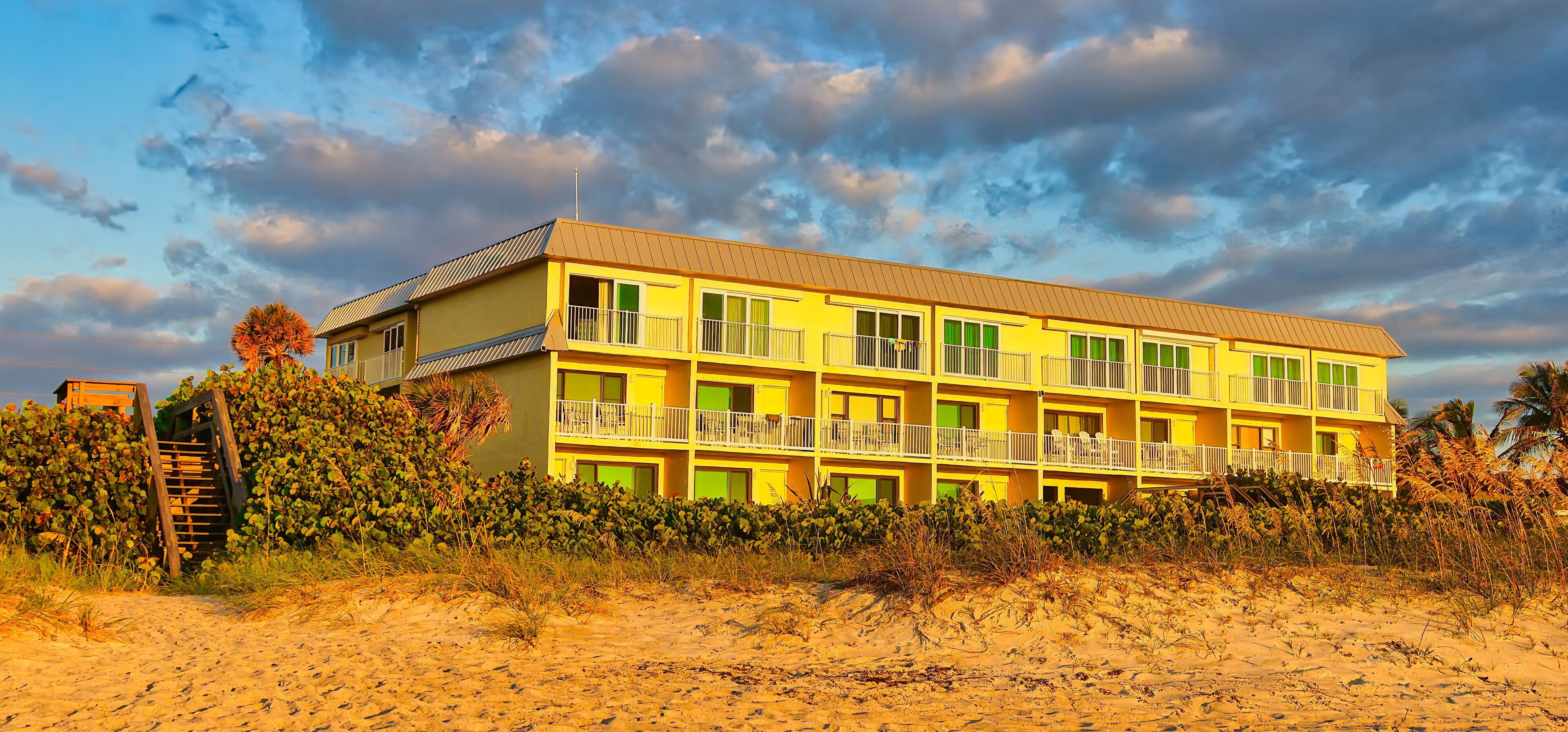 A yellow multi-story building with balconies, located on a sandy beach with vegetation.