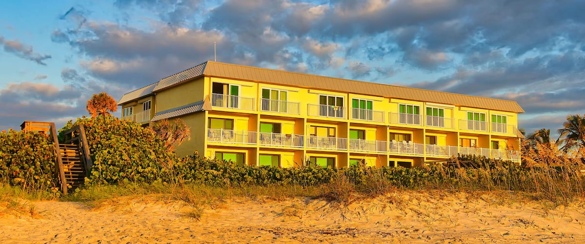 A yellow multi-story building with balconies, located on a sandy beach with vegetation.