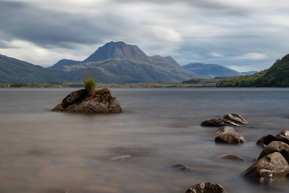 A calm lake with a rocky outcrop in the foreground and mountains in the background.