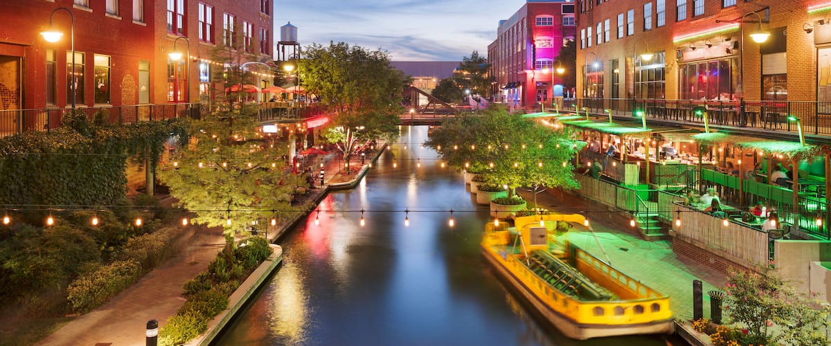 Zona urbana a orillas del río, con edificios, un barco y luces colgantes.