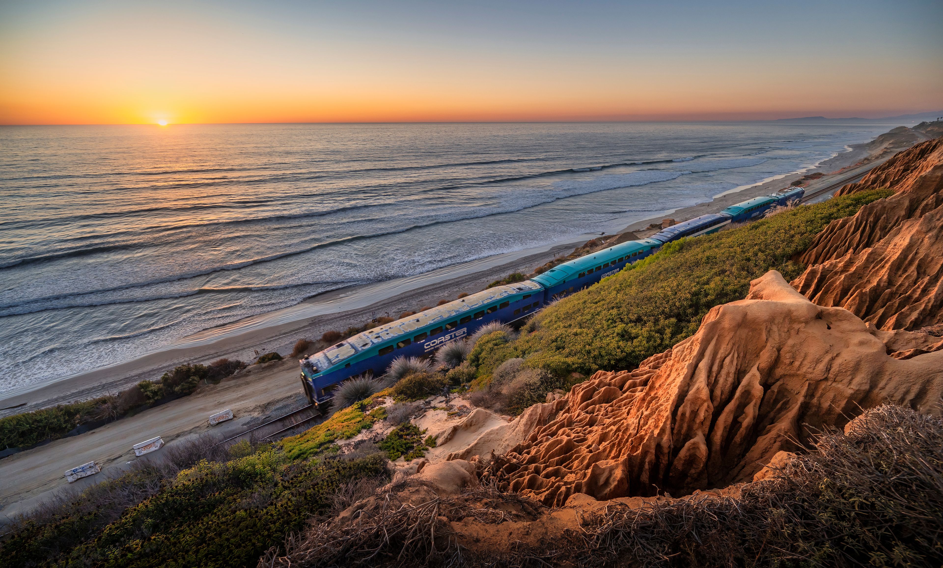 Un coucher de soleil sur la côte, avec un train sur les rails, des falaises de sable et des vagues de l’océan.
