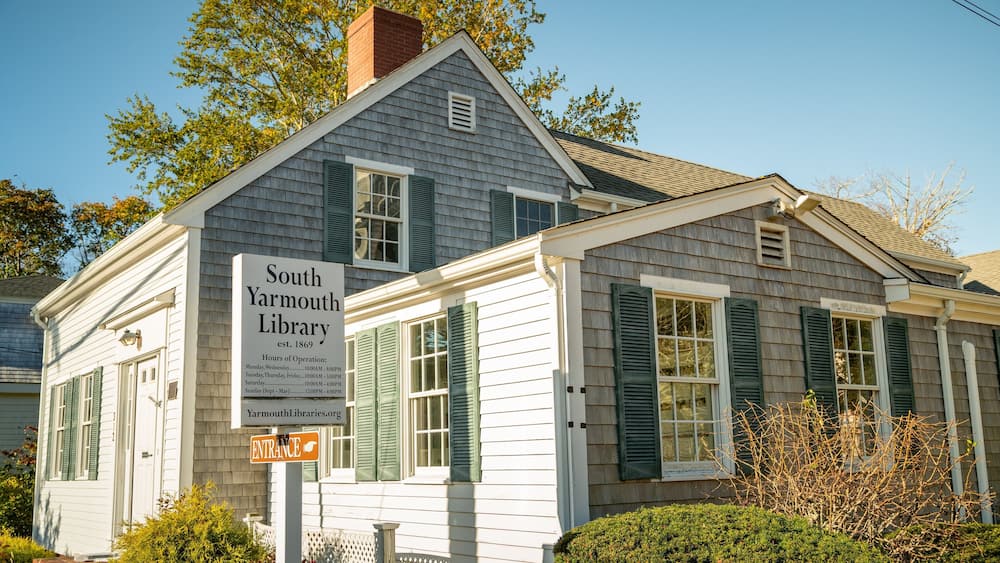 A historic South Yarmouth Library with green shutters and a sign indicating its hours of operation.