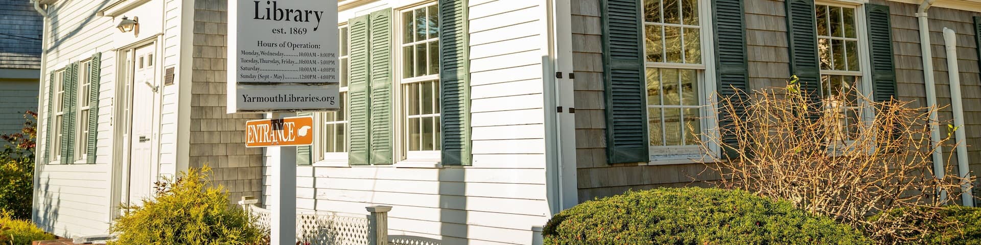 A historic South Yarmouth Library with green shutters and a sign indicating its hours of operation.