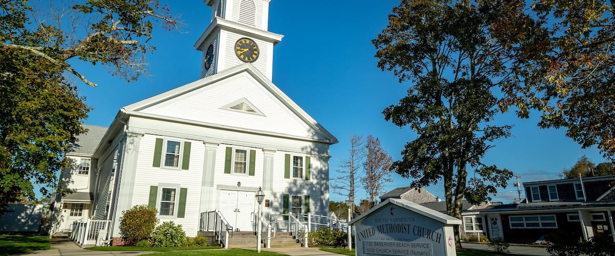 A white church with a steeple and clock, surrounded by trees and a signboard.