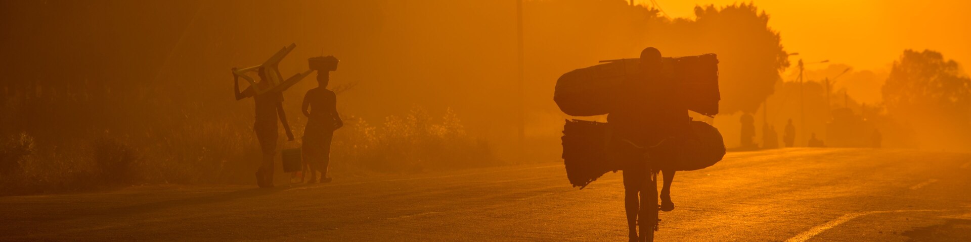 A Mozambican cyclist carrying large bags of coal to market at sunrise in the morning, along a tarmac road. Other commuters carrying items in the background. Nampula Town, Mozambique