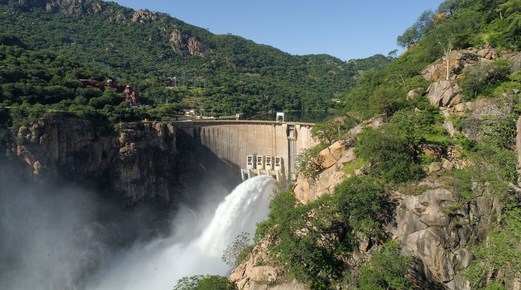 A large dam with a waterfall, surrounded by lush green hills and rocky cliffs.