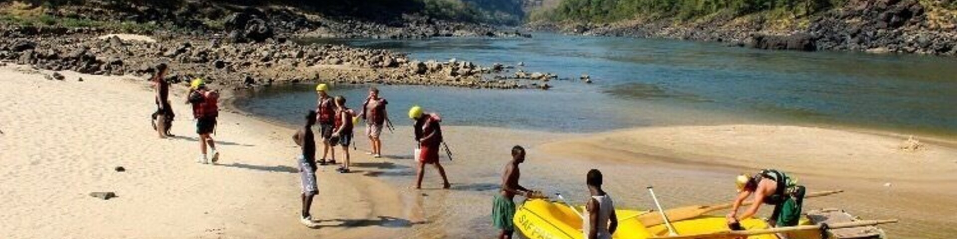 A group of people on a riverbank with boats and a rocky shoreline.