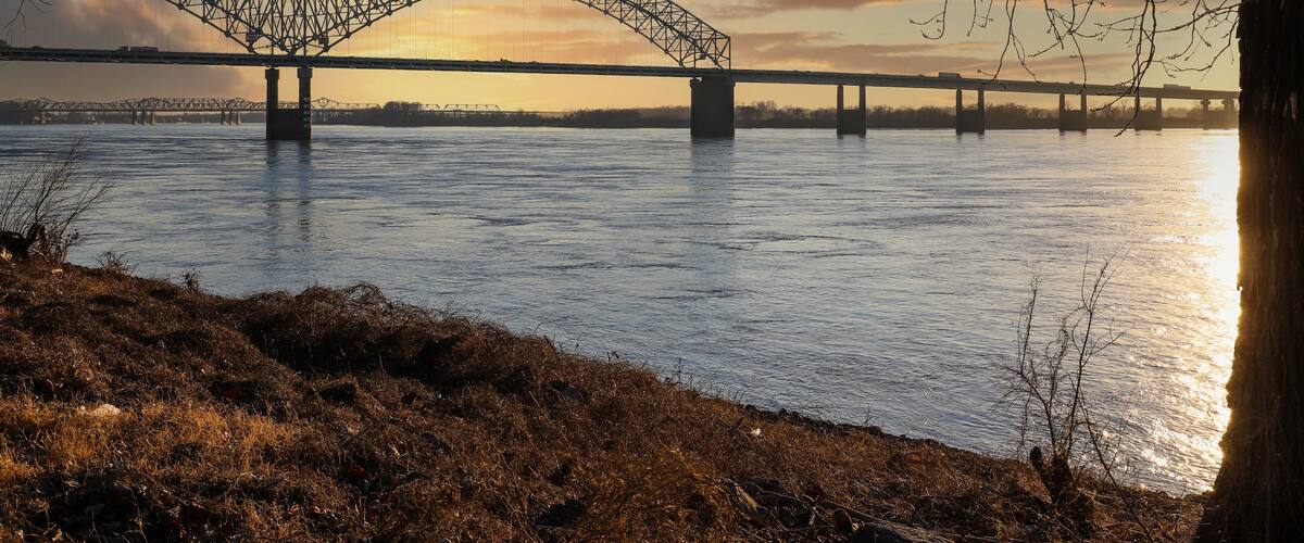 a shot of the Hernando de Soto Bridge over the flowing waters of the Mississippi river with a blue sky at sunset with bare winter trees along the banks of the river at Greenbelt Park on Mud Island