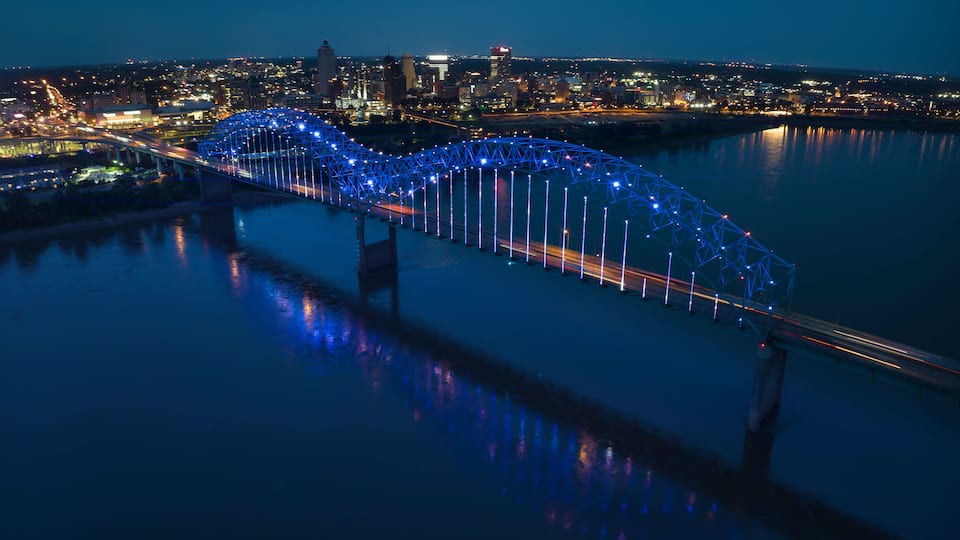 A bridge with blue lights at night, a city skyline in the background, and a river reflecting the lights.