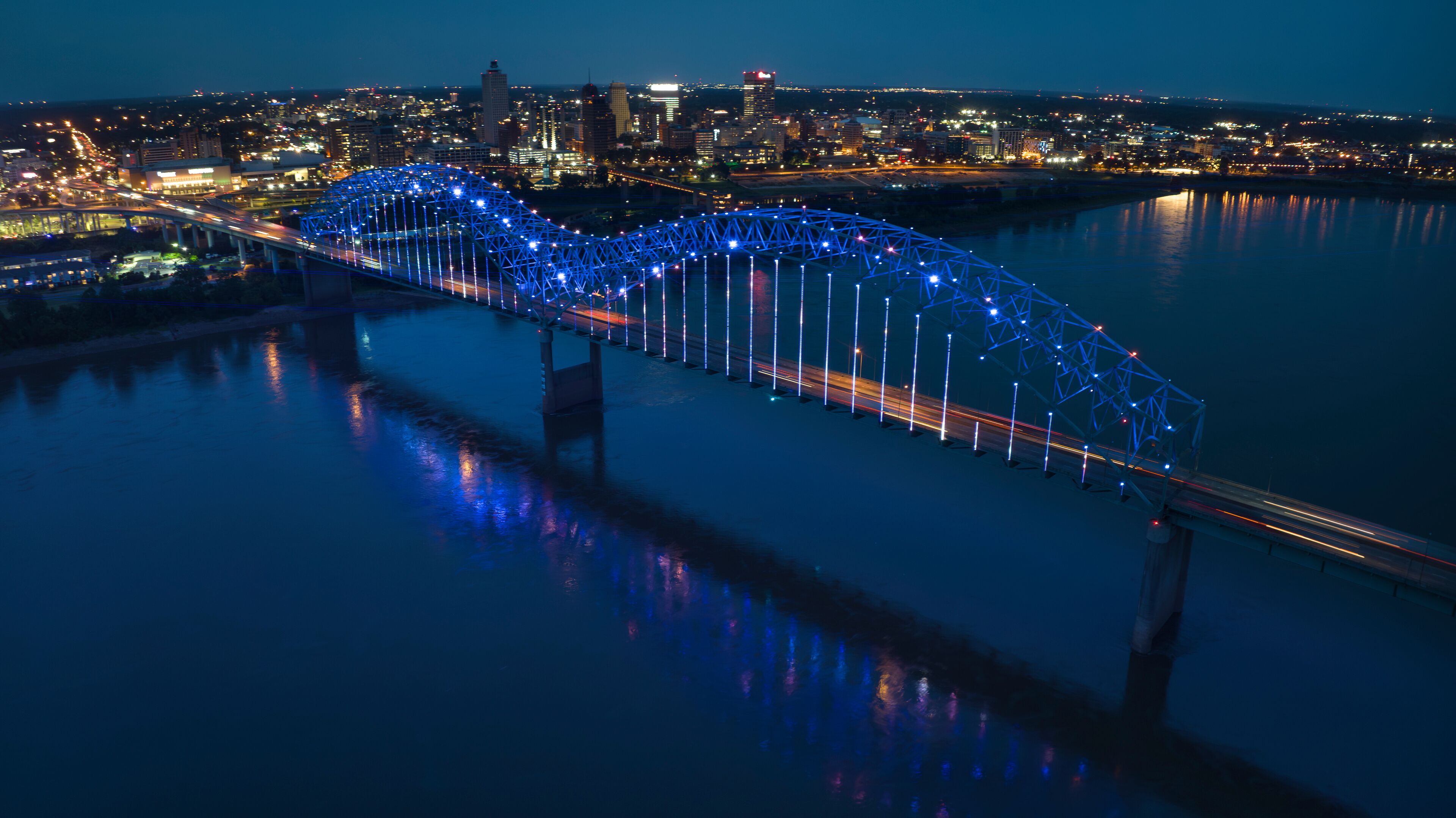 A bridge with blue lights at night, a city skyline in the background, and a river reflecting the lights.