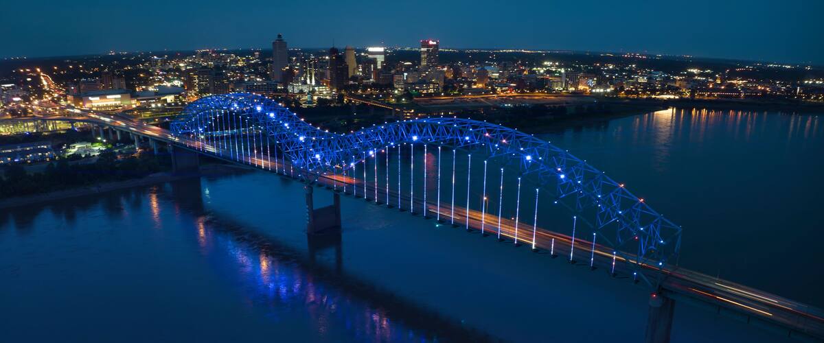 A bridge with blue lights at night, a city skyline in the background, and a river reflecting the lights.