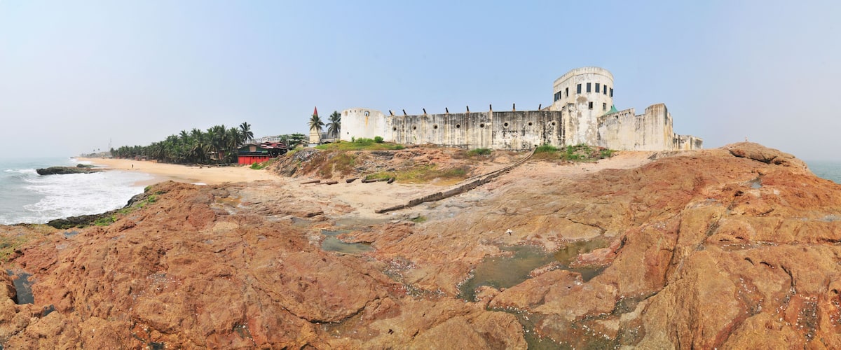 A coastal fortress with a large circular tower, rocky shoreline, and palm trees.