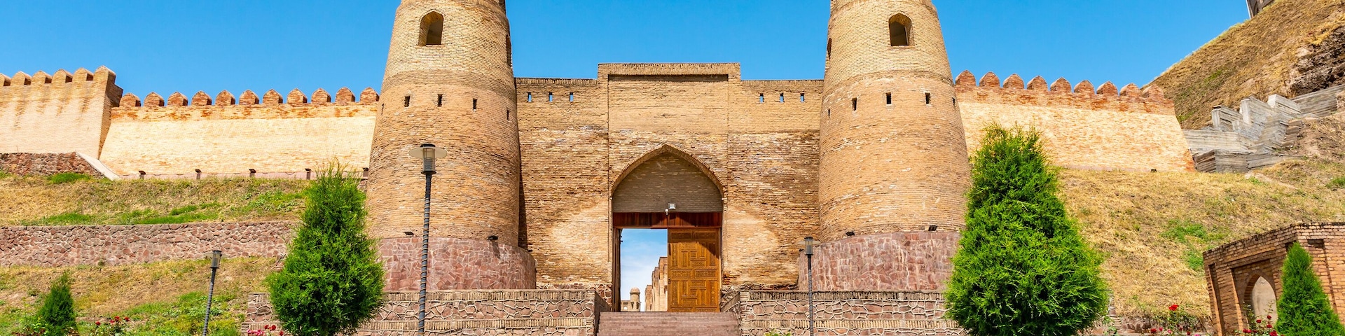 Uma fortaleza de pedra histórica com duas torres imponentes, um arco central e um caminho pavimentado que leva à entrada.