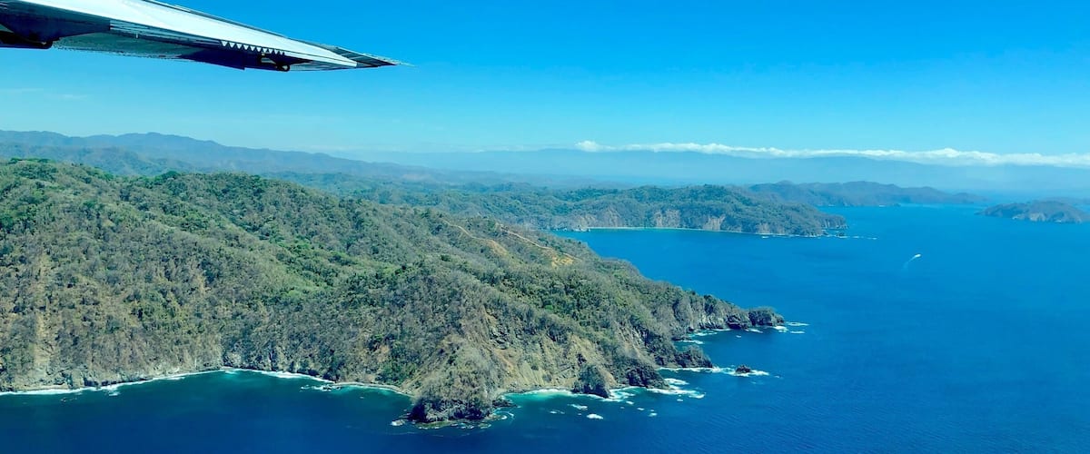 An aerial view of a coastal landscape with dense forests, rugged terrain, and a clear blue sea.
