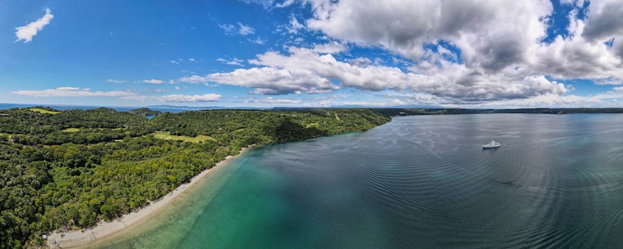 Paisagem costeira com praia, floresta densa e mar calmo com um barco.