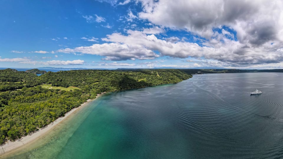 A coastal landscape with a beach, dense forest, and a calm sea with a boat.