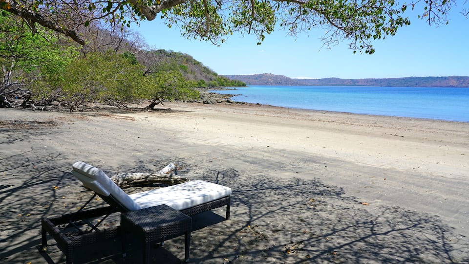 A beach with a lounge chair, trees, and a calm sea.