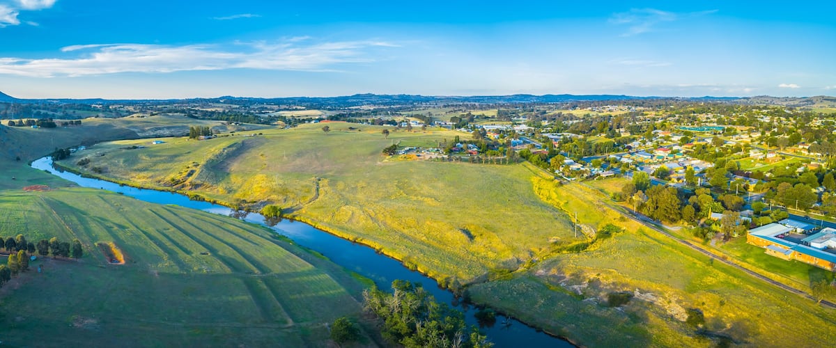 A river winding through rolling hills, a small town with houses and buildings, and distant mountains under a clear sky.