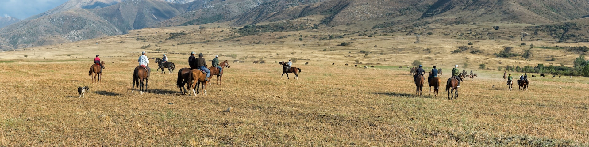 Um grupo de pessoas cavalga por um campo gramado, com montanhas ao fundo.