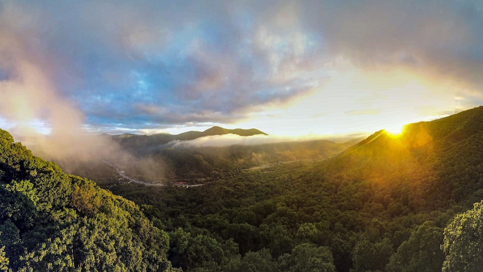 A mountainous landscape with dense forest, a winding road, and a sunrise or sunset with clouds.