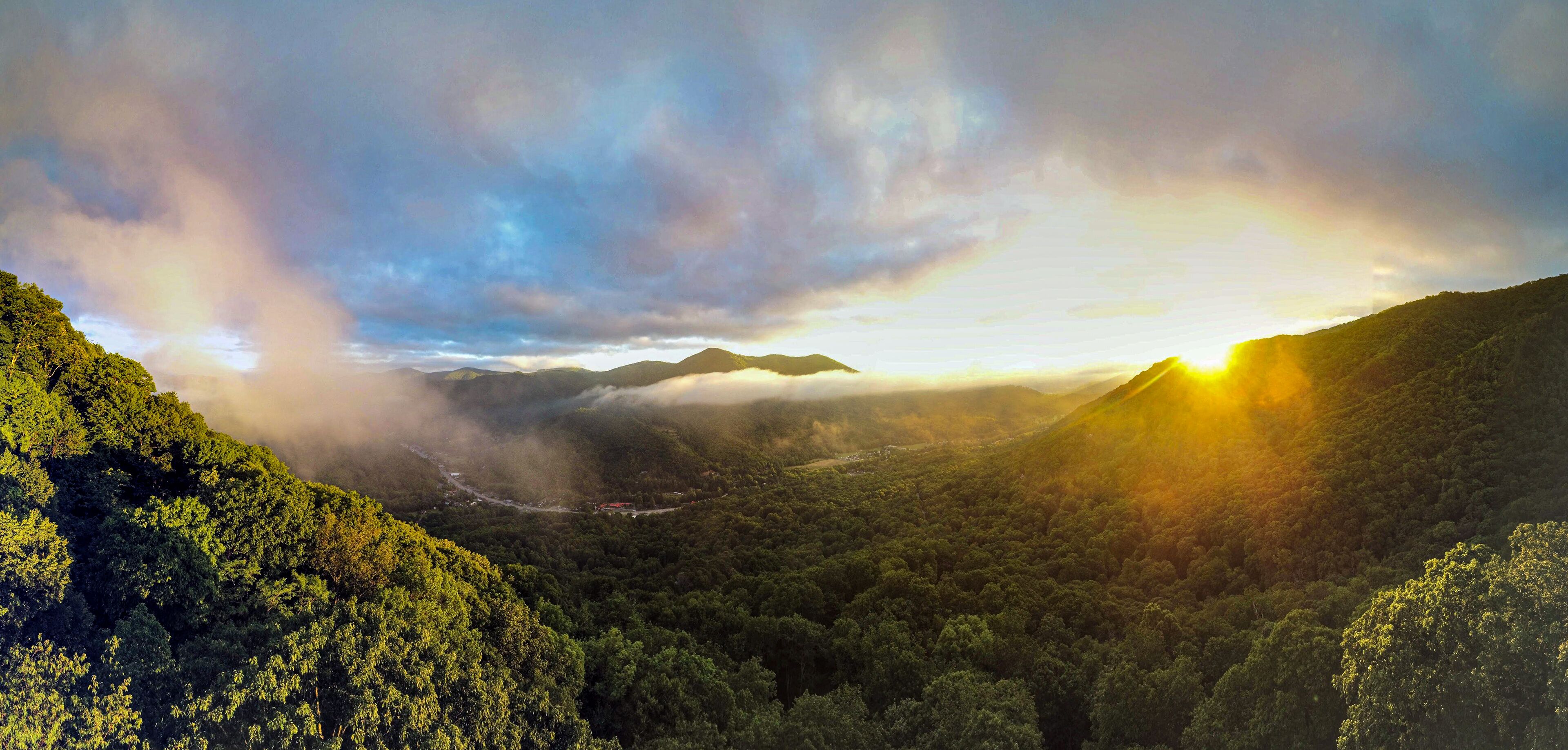 A mountainous landscape with dense forest, a winding road, and a sunrise or sunset with clouds.